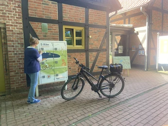 Gast vor Ladestation Eine Person steht vor einer E-Bike-Ladestation auf einem gepflasterten Weg neben einem Gebäude.