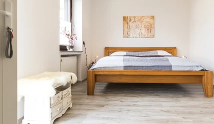 Helles Schlafzimmer mit Holzbett, weißem Nachttisch und Wandgemälde, Fenster auf der linken Seite.
