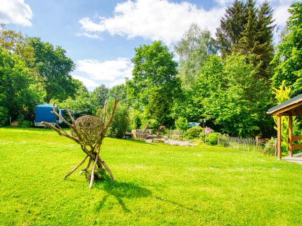 Garten.jpeg Weitläufiger, grüner Garten mit Bäumen, einer Holzterrasse und einer dekorativen Astskulptur.