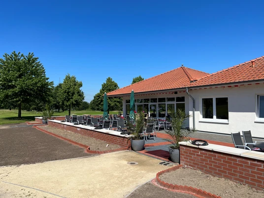 Außenterrasse eines Restaurants am Golfplatz mit roten Ziegeln und Sonnenschirmen unter blauem Himmel.