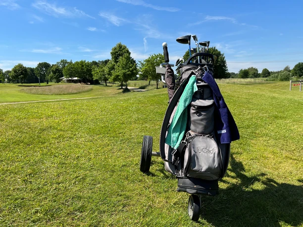Golfplatz Rehburg-Loccum Caddy A golf trolley stands on the lush green of a golf course, surrounded by trees and blue sky.