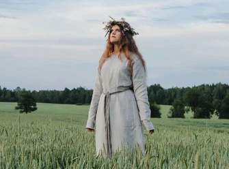 Midsommer Eine Frau trägt ein langes, helles Kleid und Blumenkranz, steht in einem grünen Weizenfeld.