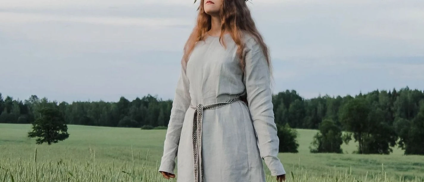 Midsommer Eine Frau trägt ein langes, helles Kleid und Blumenkranz, steht in einem grünen Weizenfeld.