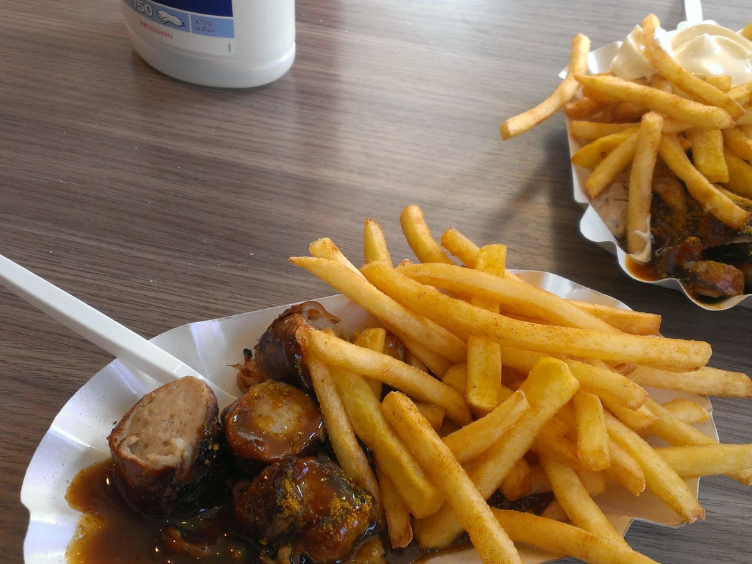 Currywurst Pommes Ein Teller mit Currywurst, dazu knusprige Pommes und eine Gabel auf einem Holztisch drapiert.