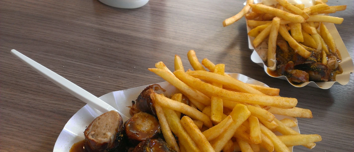 Currywurst Pommes Ein Teller mit Currywurst, dazu knusprige Pommes und eine Gabel auf einem Holztisch drapiert.