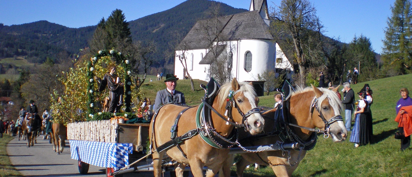 Leonhardiritt Unterammergau Zwei Pferde ziehen einen geschmückten Wagen beim Leonhardiritt in Unterammergau vorbei an einer Kirche.