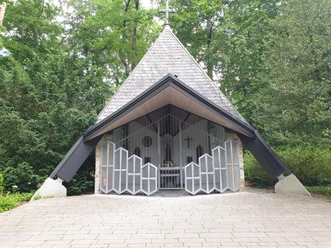 Kapelle mit schrägem Dach in einem Waldgebiet, umgeben von hohen Bäumen, mit offenem Eingang.