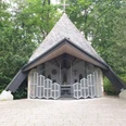 Kapelle mit schrägem Dach in einem Waldgebiet, umgeben von hohen Bäumen, mit offenem Eingang.