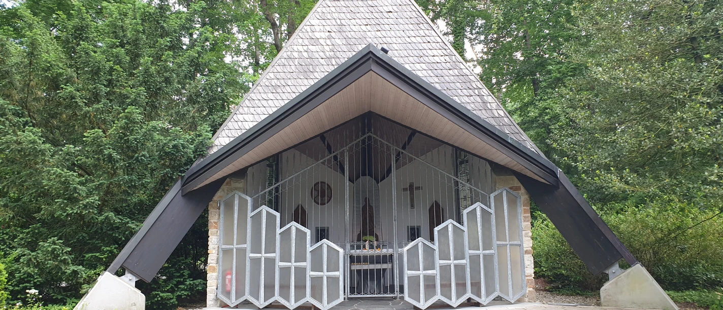 Kapelle mit schrägem Dach in einem Waldgebiet, umgeben von hohen Bäumen, mit offenem Eingang.