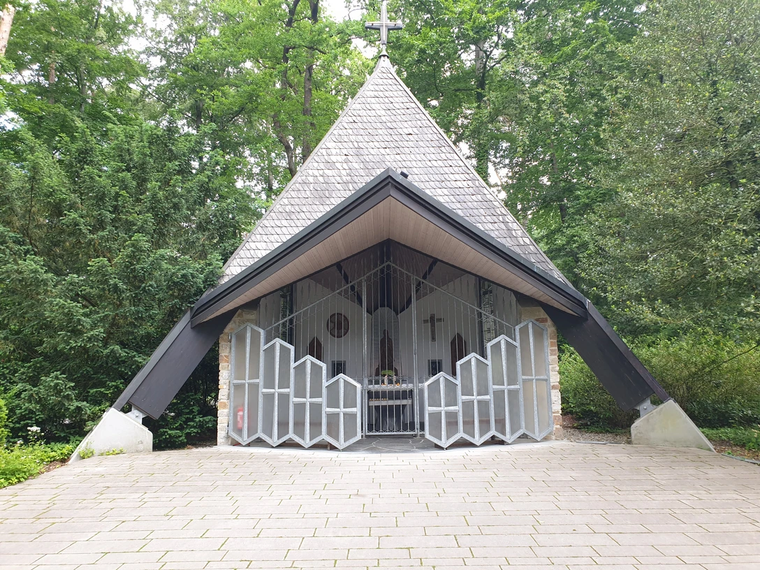 Friedenskapelle.jpg Kapelle mit schrägem Dach in einem Waldgebiet, umgeben von hohen Bäumen, mit offenem Eingang.