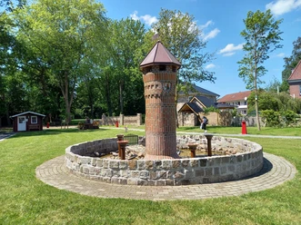 Mehrgenerationenplatz "Lüttje Festung" in Apen Der Mehrgenerationenplatz "Lüttje Festung" in Apen zeigt einen zentralen Backsteinturm inmitten einer kreisförmigen Steinmauer, umgeben von gepflegtem Rasen und Bäumen.