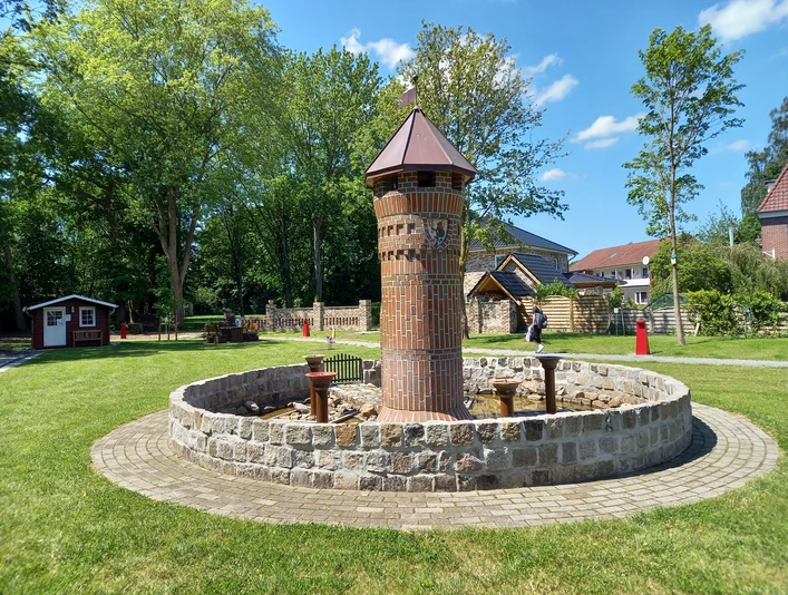 Mehrgenerationenplatz "Lüttje Festung" in Apen Der Mehrgenerationenplatz "Lüttje Festung" in Apen zeigt einen zentralen Backsteinturm inmitten einer kreisförmigen Steinmauer, umgeben von gepflegtem Rasen und Bäumen.