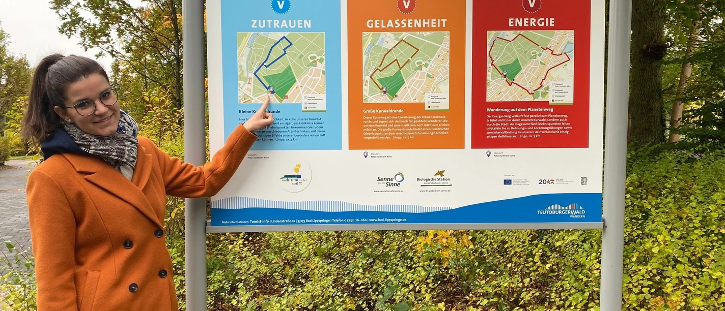 2021-10-22_Erlebbare VitalWanderWelt im Heilwald.jpg Frau zeigt auf VitalWanderWelt-Infotafel im Heilwald, die Wanderwege und Themen erklärt.