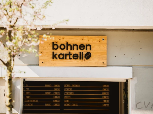 Bohnen Kartell in Rüttenscheid, Essen <p>Holzschild "Bohnen Kartell" über einer modernen Fassade, Baum mit zarten Blüten im Vordergrund.</p>