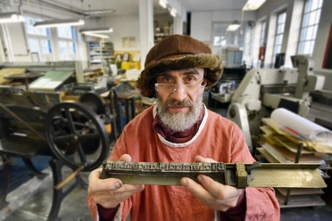 Die Buchmacherey in Velbert-Langenberg mit Hans Josef Altmann.jpg Ein Buchdrucker in traditioneller Kleidung hält eine Druckplatte in einer historischen Werkstatt.