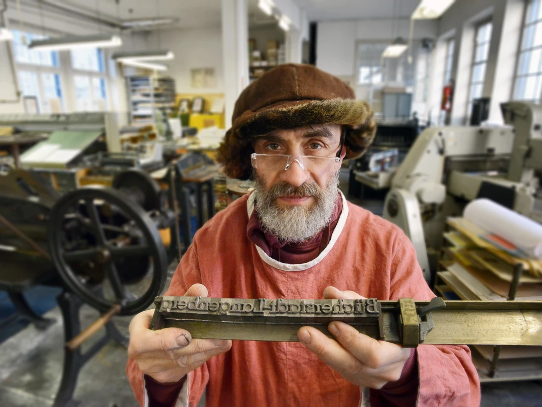 Die Buchmacherey in Velbert-Langenberg mit Hans Josef Altmann.jpg Ein Buchdrucker in traditioneller Kleidung hält eine Druckplatte in einer historischen Werkstatt.