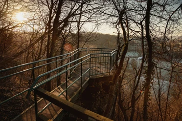 Metallplattform mit seitlichen Geländern in einem Waldgebiet, bietet Blick auf einen See bei Sonnenuntergang.
