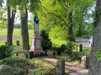 Marienstatue im Grünen, umgeben von Weg, Bäumen und Kerzen, neben landwirtschaftlich genutzter Fläche.