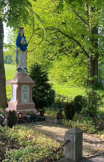 Marienstatue im Grünen, umgeben von Weg, Bäumen und Kerzen, neben landwirtschaftlich genutzter Fläche.