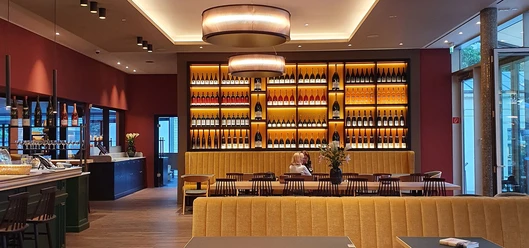 Elisenbrunnen Gastronomie Innenraum mit langen Tischen, an denen viele Stühle stehen, und eine Sitzbank im Vordergrund. Beleuchtetes Weinregal an der Wand mit vielen verschiedenen Flaschen.