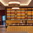 Elisenbrunnen Gastronomie Innenraum mit langen Tischen, an denen viele Stühle stehen, und eine Sitzbank im Vordergrund. Beleuchtetes Weinregal an der Wand mit vielen verschiedenen Flaschen.