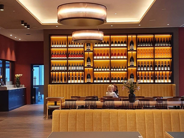 Elisenbrunnen Gastronomie Innenraum mit langen Tischen, an denen viele Stühle stehen, und eine Sitzbank im Vordergrund. Beleuchtetes Weinregal an der Wand mit vielen verschiedenen Flaschen.