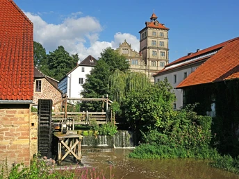 Schloss Brake Ölmühle Historisches Schloss mit Mühle und Wasserrad, umgeben von grüner Vegetation und Gebäuden.