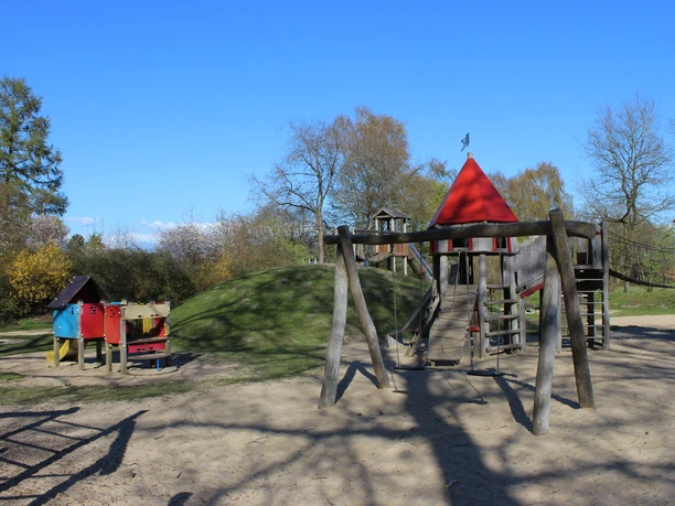 Spielplatz Cadenberge Holzspielplatz in Cadenberge mit Rutsche, Schaukeln und roten Türmen, umgeben von grünen Bäumen unter blauem Himmel.