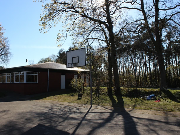 Spielplatz Cadenberge Basketballplatz in Cadenberge mit Korb neben rotem Backsteingebäude, umgeben von Bäumen im Sonnenschein.
