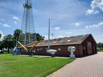 Rotes Backsteingebäude mit Freifläche; historische Windturbinen; teils bewölkter Himmel.
