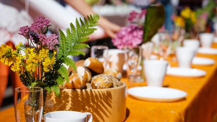 Ein langer Tisch mit Blumen, Brötchen und Geschirr, bei Sonnenlicht für ein gemeinschaftliches Frühstück.