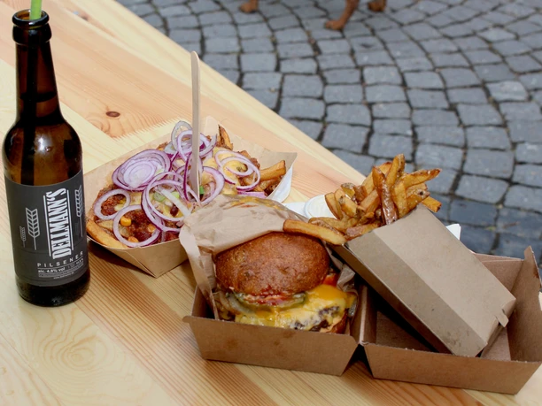 Bergischer Feierabendmarkt Ein Gourmet-Burger mit Pommes, Zwiebelringen und einer Flasche Bier auf einem Holztisch im Freien.