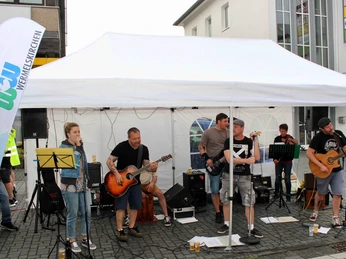 Bergischer Feierabendmarkt Straßenband spielt unter weißem Zelt, mehrere Musiker mit Gitarren und Mikrofonen auf einem Gehweg.