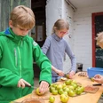 Kinder-Aktion auf Gut Halfeshof in Mettmann Drei Kinder schälen Äpfel an einem Holztisch vor einem Hintergrund aus grünen Brettern und Backsteinwänden.