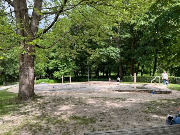 Kinderspielplatz mit Wasserflächen und Sand, umgeben von grünem Laubwald und schattenspendenden Bäumen.