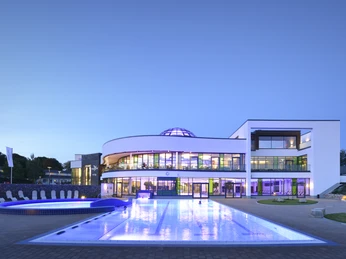 Aussenbereich der carpesol SPA Therme Moderne Gebäude mit beleuchtetem Pool und gläserner Kuppel vor Abendhimmel.Modern building with illuminated pool and glass dome against the evening sky.Moderne bygning med oplyst pool og glaskuppel mod aftenhimlen.Modern gebouw met verlicht zwembad en glazen koepel tegen de avondlucht.