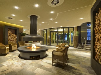 Entspannungsbereiche in der carpesol SPA Therme Gemütlicher Loungebereich mit zentralem Kamin, umgeben von Steinsitzen und Holzstapeln.Cozy lounge area with central fireplace, surrounded by stone seats and stacks of wood.Hyggeligt loungeområde med central pejs, omgivet af stensæder og bunker af træ.Gezellige zithoek met centrale open haard, omringd door stenen zitjes en stapels hout.