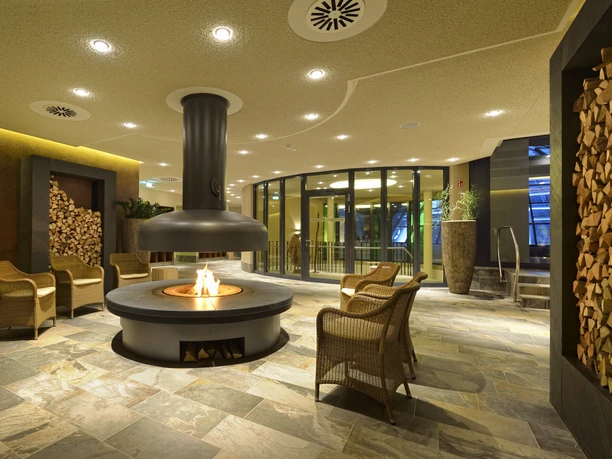 Entspannungsbereiche in der carpesol SPA Therme Cozy lounge area with central fireplace, surrounded by stone seats and stacks of wood.