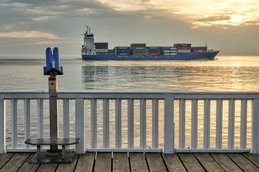 Cuxland, Cuxhaven - Alte Liebe Ein Containerschiff fährt bei Sonnenuntergang vor einer blauen Aussichtsplattform auf dem Wasser.