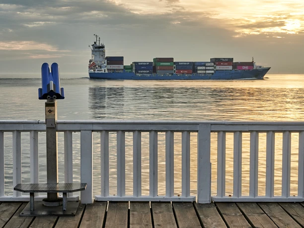 Cuxland, Cuxhaven - Alte Liebe Ein Containerschiff fährt bei Sonnenuntergang vor einer blauen Aussichtsplattform auf dem Wasser.