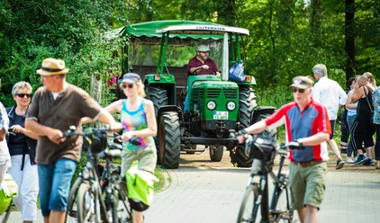 © SOS-Hof Bockum Philipp Schulze.jpg Menschen gehen oder fahren Fahrrad bei einem Fest auf dem Hof. Ein grüner Traktor steht auf dem Weg. Eine Person fährt den Traktor. Viele Menschen tragen Sommerkleidung. Die Sonne scheint. Es wirkt fröhlich und lebendig.