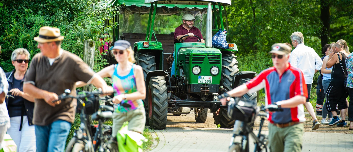 © SOS-Hof Bockum Philipp Schulze.jpg Menschen gehen oder fahren Fahrrad bei einem Fest auf dem Hof. Ein grüner Traktor steht auf dem Weg. Eine Person fährt den Traktor. Viele Menschen tragen Sommerkleidung. Die Sonne scheint. Es wirkt fröhlich und lebendig.