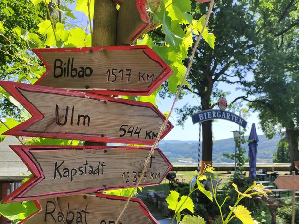 1655288876960.jpg Holzwegweiser im Grünen mit Entfernungen zu Städten neben einem Biergarteneingang bei Sonnenschein.