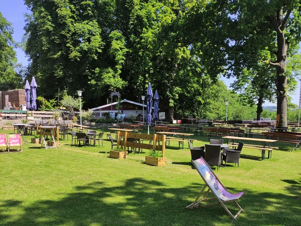 Grüner Biergarten mit Holzstühlen und Liegestühlen unter hohen Bäumen bei sonnigem Wetter.