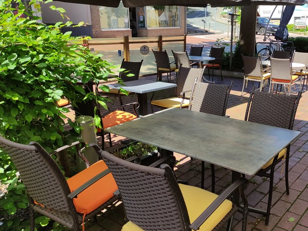 Freiluftterrasse mit Tischen und Stühlen aus Korbgeflecht, umgeben von grünen Pflanzen und Ziegelboden.