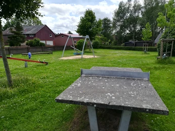 Spielplatz Cadenberge OT Geversdorf Grüner Spielplatz mit Wippe, Schaukeln und Tischtennisplatte, umgeben von Bäumen und Häusern.
