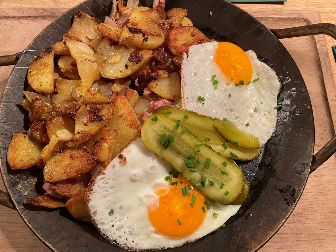 Gebratene Kartoffelscheiben mit knusprigem Speck, Spiegeleiern und Gurkenscheiben auf dunklem Teller.Fried potato slices with crispy bacon, fried eggs and cucumber slices on a dark plate.Stegte kartoffelskiver med sprød bacon, spejlæg og agurkeskiver på en mørk tallerken.Gebakken aardappelschijfjes met knapperig spek, gebakken eieren en komkommerschijfjes op een donker bord.