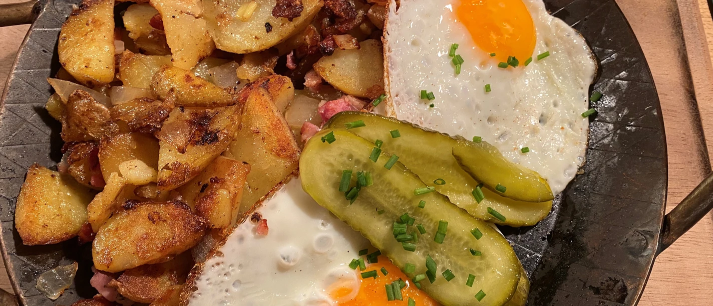 Bratkartoffeln Gebratene Kartoffelscheiben mit knusprigem Speck, Spiegeleiern und Gurkenscheiben auf dunklem Teller.