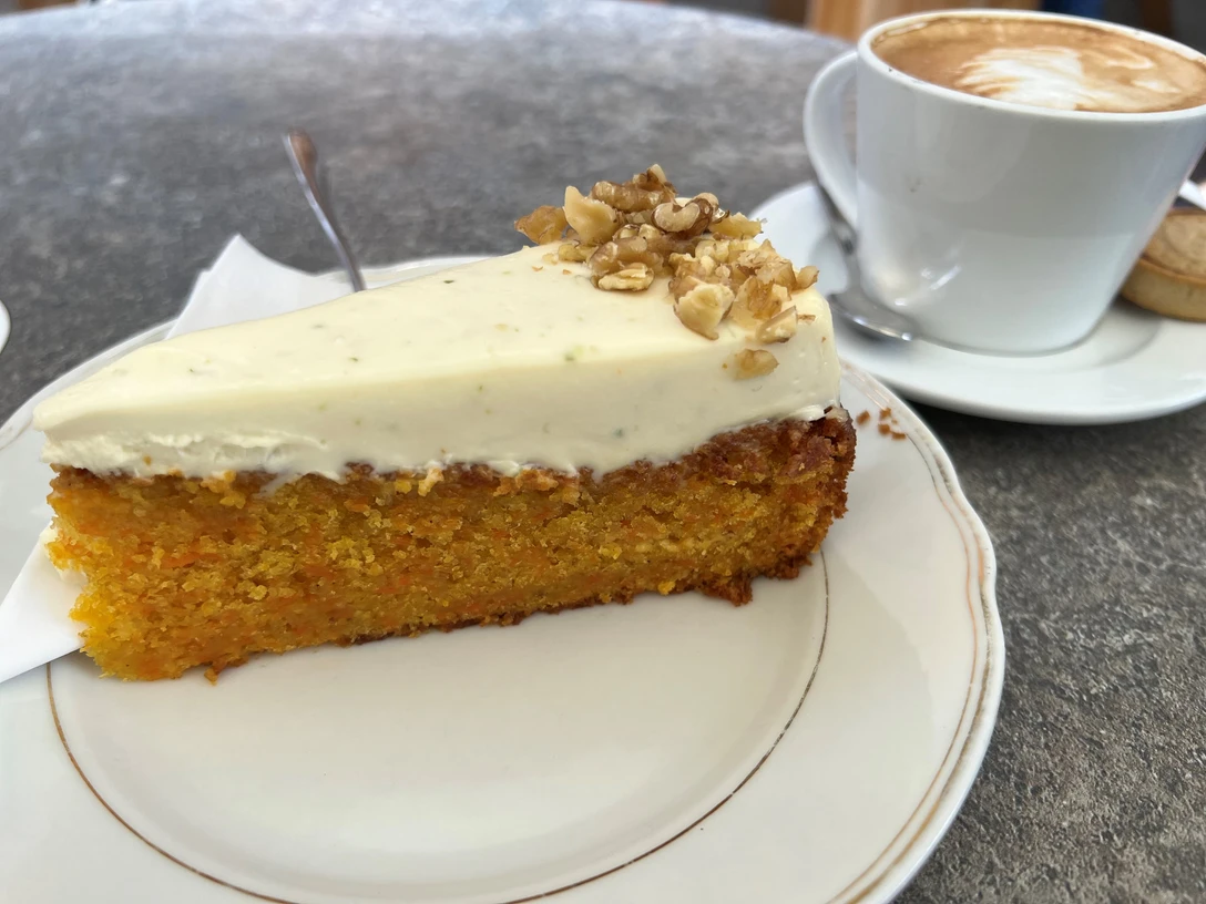 Ein Stück Karottenkuchen gekrönt mit Frischkäse und Nüssen auf einem Teller neben einer Kaffeetasse.A slice of carrot cake topped with cream cheese and nuts on a plate next to a coffee cup.Et stykke gulerodskage toppet med flødeost og nødder på en tallerken ved siden af en kaffekop.Een plak worteltaart met roomkaas en noten op een bord naast een koffiekopje.