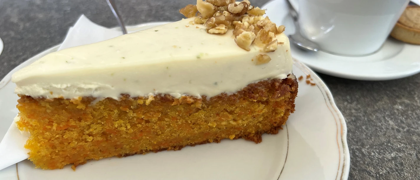 Een plak worteltaart met roomkaas en noten op een bord naast een koffiekopje.